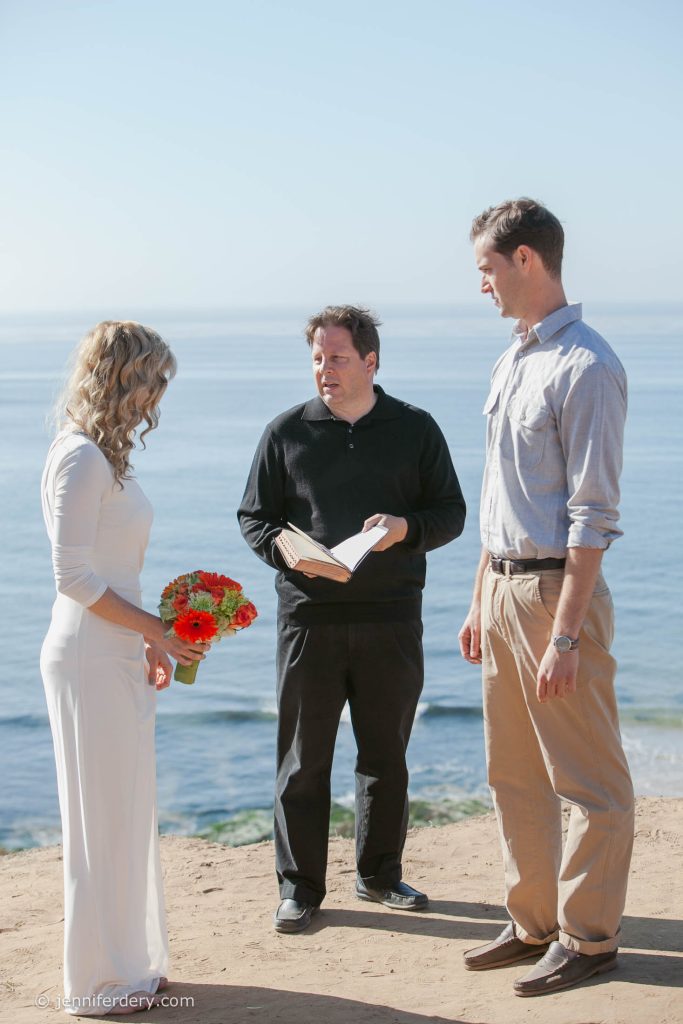 A bride in a white dress holding a bouquet, a groom in a light shirt and khaki pants, and an officiant with an open book stand on a sandy cliff overlooking the ocean under a clear sky.