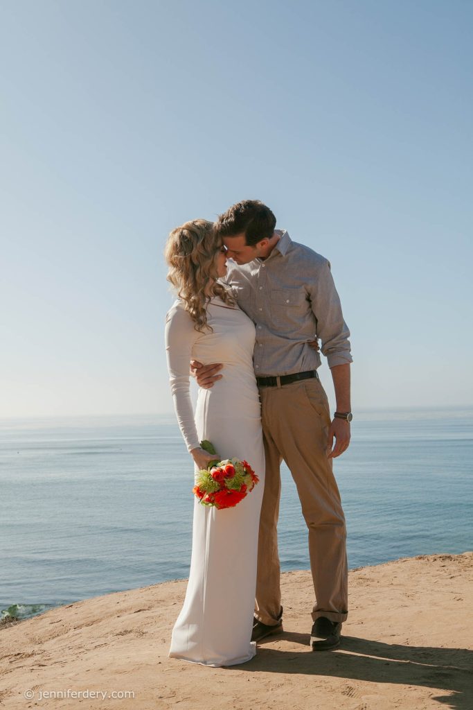 A couple stands on a cliff overlooking the ocean, sharing a kiss. The woman wears a long white dress and holds a bouquet of flowers, while the man wears a gray shirt and khaki pants. The sky is clear and blue.