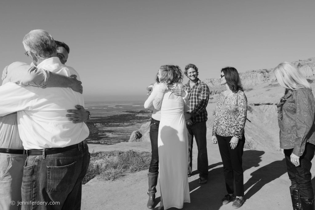 A group of people stand on a scenic cliffside overlooking the ocean. Two pairs of people are hugging while others look on, smiling. The scene appears joyful and relaxed under a clear sky.