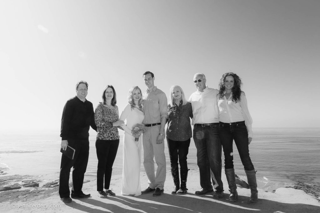 A group of seven people, including a bride in a white dress holding a bouquet, stand closely together and smile at the camera on a cliffside overlooking the ocean. The photo is in black and white.