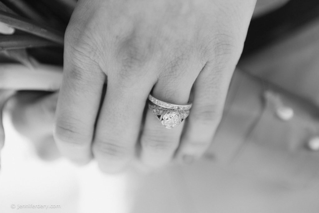 A close-up, black-and-white image of a hand wearing a diamond engagement ring and wedding band, gently resting on a bouquet of flowers.