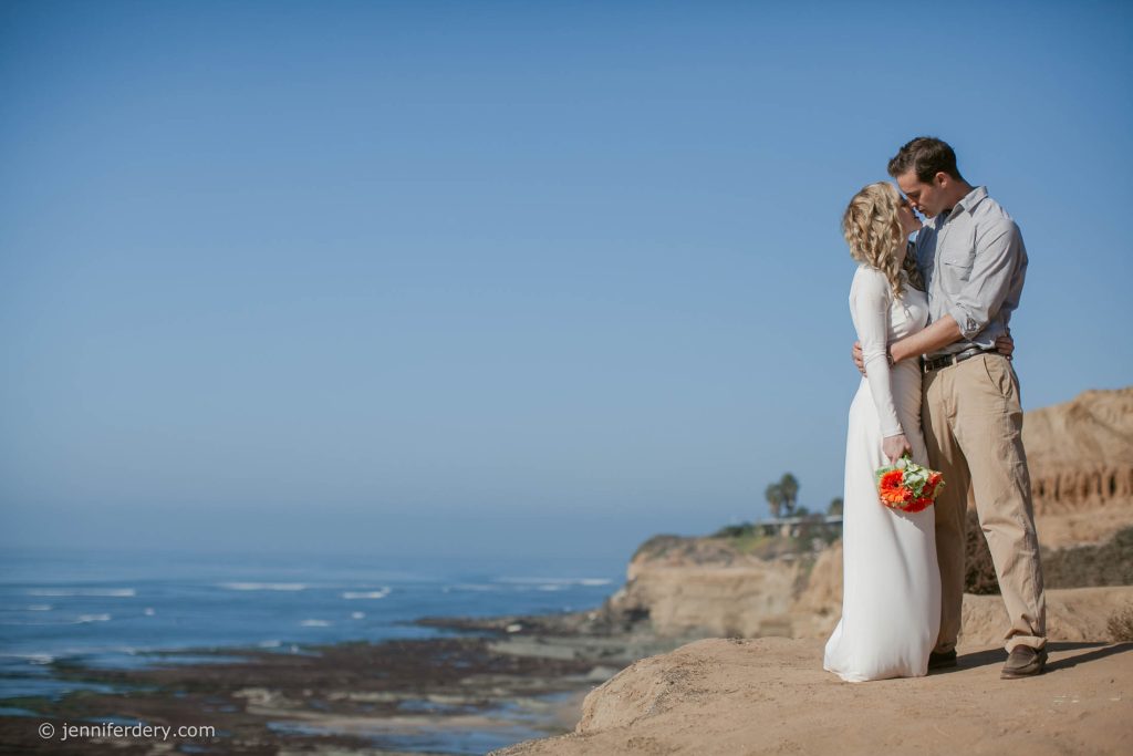 A bride and groom embrace on a cliff overlooking the ocean. The bride wears a white dress and holds a bouquet of flowers, while the groom is in a light shirt and khakis. The sky is clear and the coastline stretches in the background.