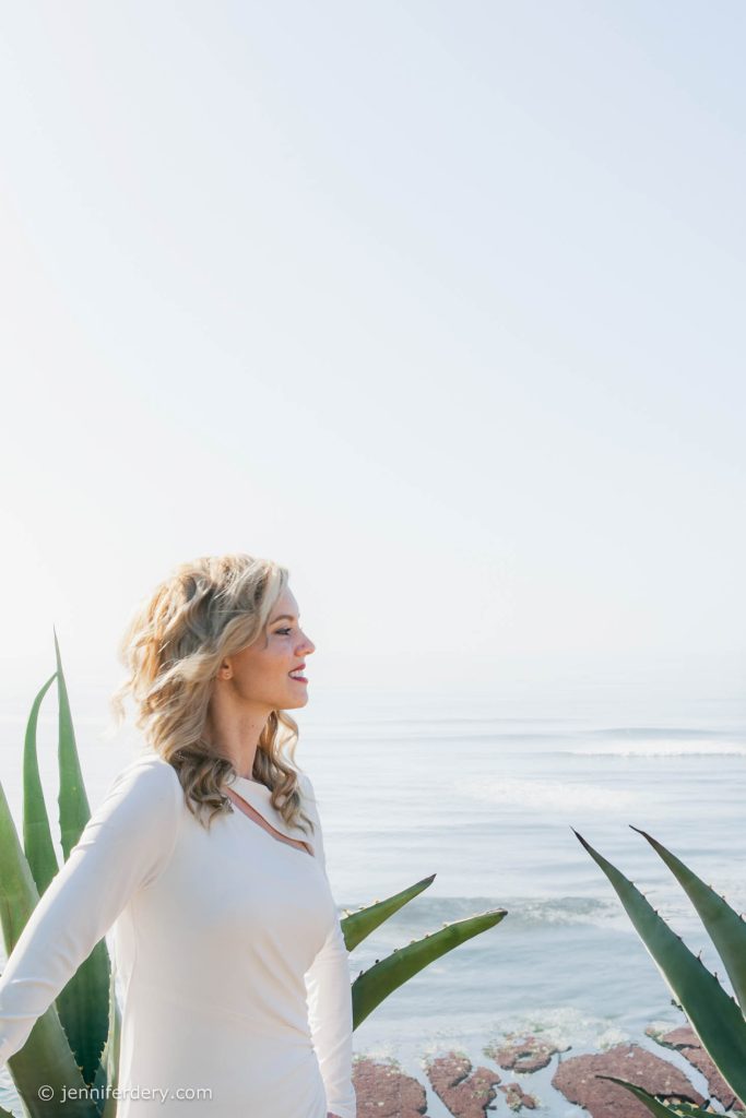 A woman in a white dress stands near green plants, looking out over a calm ocean. The scene is bright and serene, with soft sunlight illuminating the water and sky in the background.