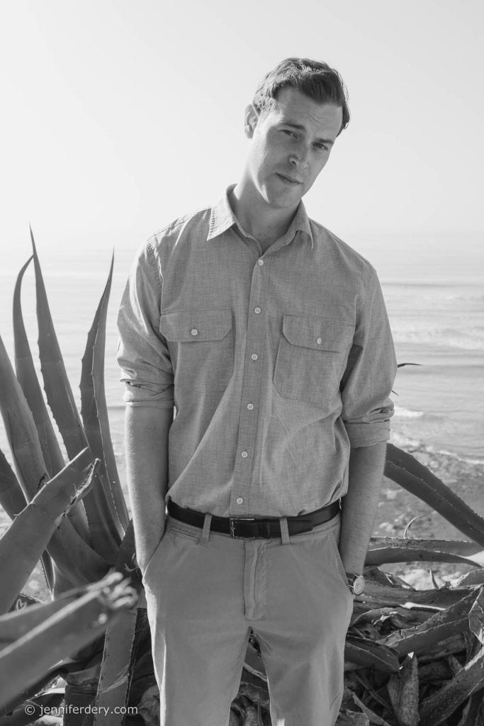 A young man in a button-up shirt and pants stands near large agave plants by the ocean, looking slightly pensive, with his hands in his pockets. The image is in black and white.