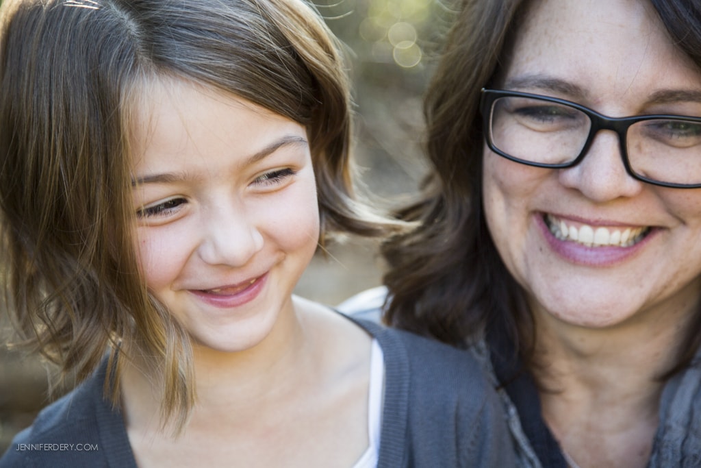 A smiling woman with glasses and a young girl with short hair are close together outdoors. The girl is looking down, and the woman is looking forward, both appearing joyful.