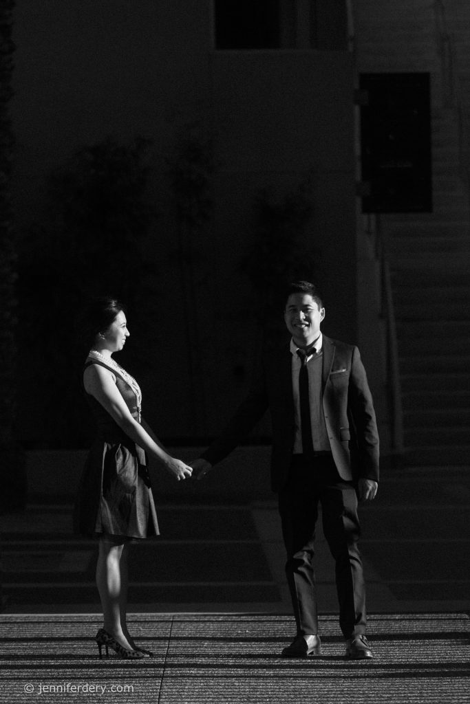 A woman and man dressed formally stand holding hands on a patterned ground, facing each other and smiling, illuminated by a spotlight against a dark background.