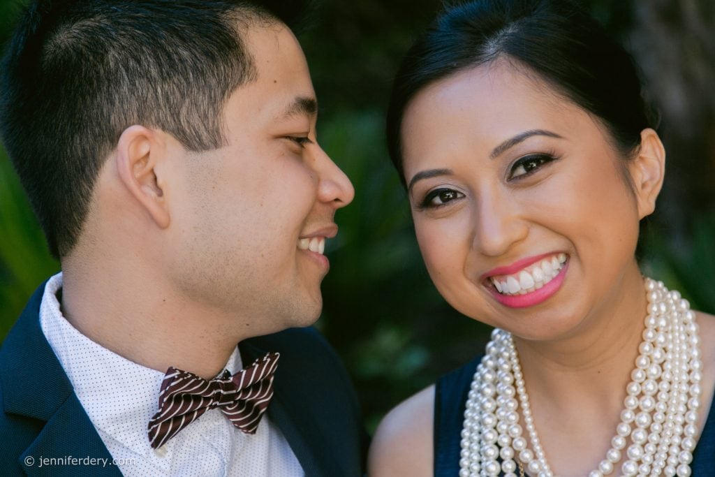 A smiling woman wearing a pearl necklace and bright pink lipstick poses next to a man in a suit and striped bow tie, who is looking at her and smiling. They appear happy and are outdoors with greenery in the background.