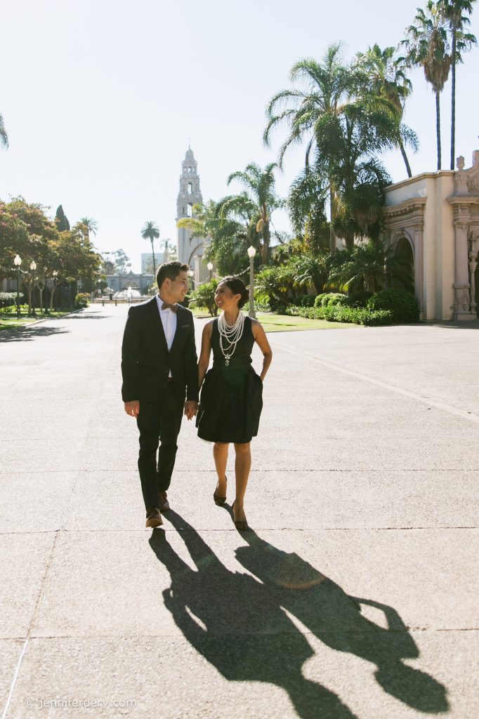 A well-dressed couple walks hand in hand along a sunlit, palm-lined path in a park, casting long shadows on the pavement. Ornate buildings and a tall tower are visible in the background.