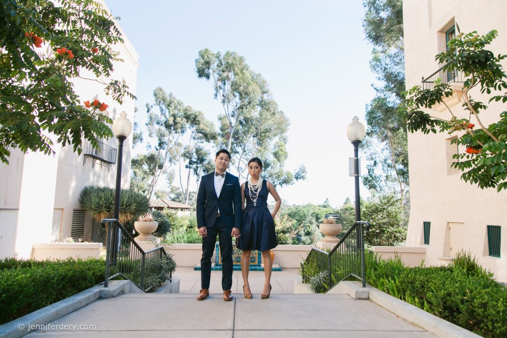 A man in a suit and a woman in a dark dress stand side by side on a walkway between two buildings, surrounded by greenery and trees on a sunny day.