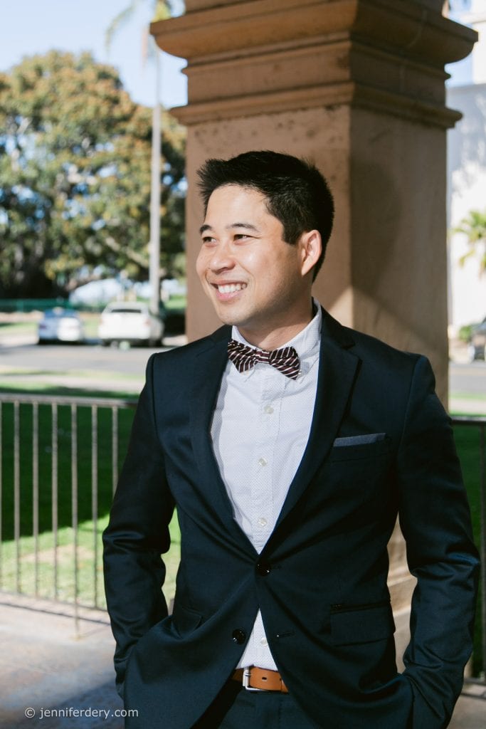 A man in a dark suit and bow tie stands outdoors, smiling and looking to the side. Sunlight highlights his face, and greenery and parked cars are visible in the background.
