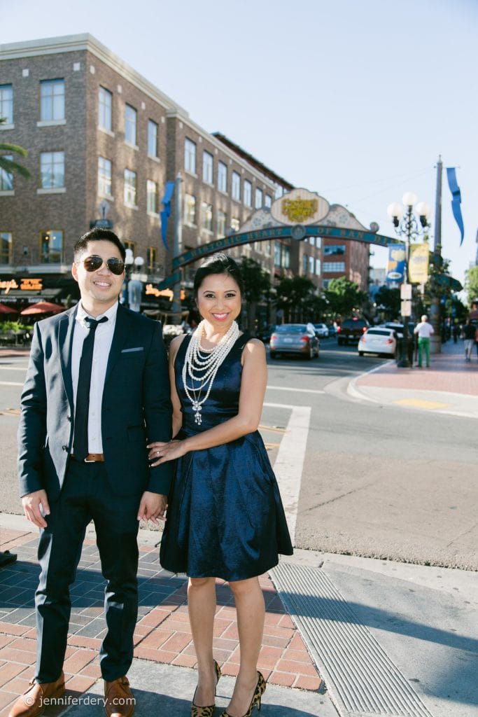 A smiling couple dressed formally stands on a city street corner in daylight, with the Gaslamp Quarter arch and buildings visible in the background. The woman wears a navy dress and pearls; the man wears a dark suit and sunglasses.
