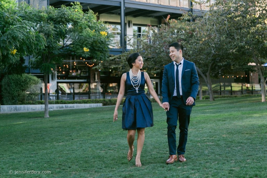 A smiling couple dressed in formal attire walks hand-in-hand across a green lawn, with trees and a modern building in the background.