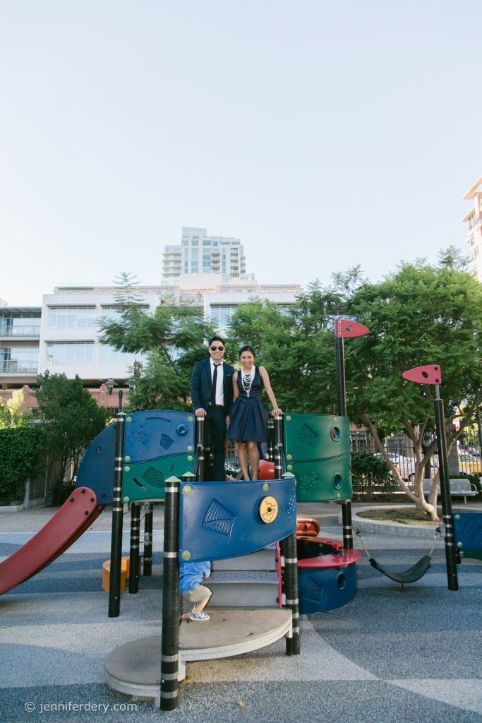 A man and woman dressed in formal clothing stand on top of colorful playground equipment, smiling at the camera. Trees and apartment buildings are visible in the background. A child is partially visible below them.
