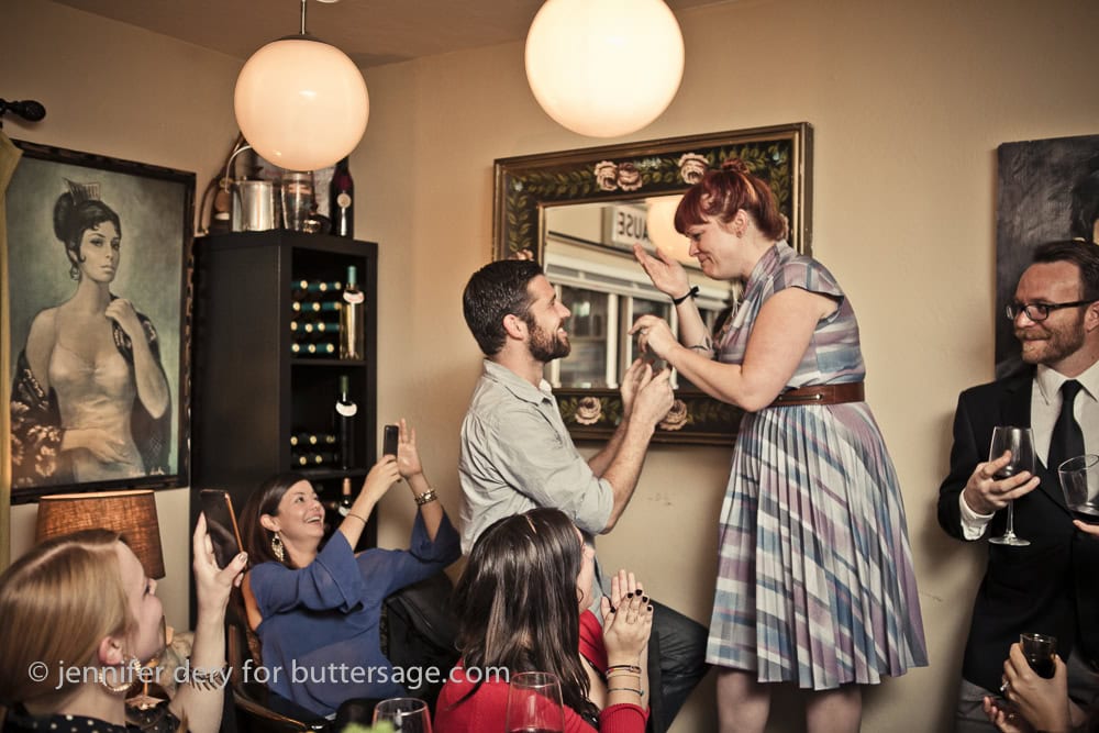 A man kneels and proposes to a woman standing on a chair as friends cheer and clap. The warmly lit room features a wine rack, large painting, and mirror, adding charm to the celebration.