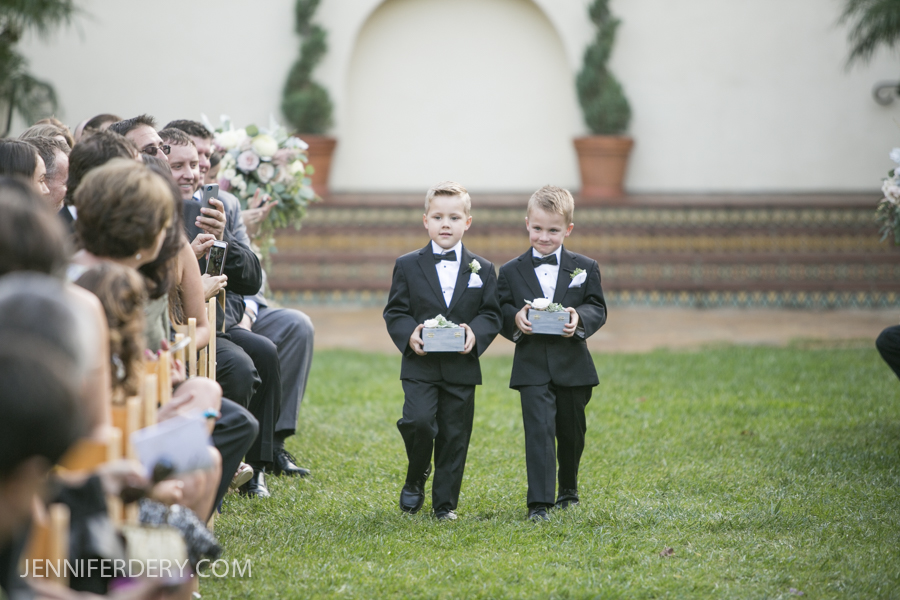 Two young boys in black tuxedos walk down an outdoor wedding aisle, each holding a small box. Guests seated on either side watch and take photos. The background features greenery and decorative steps.
