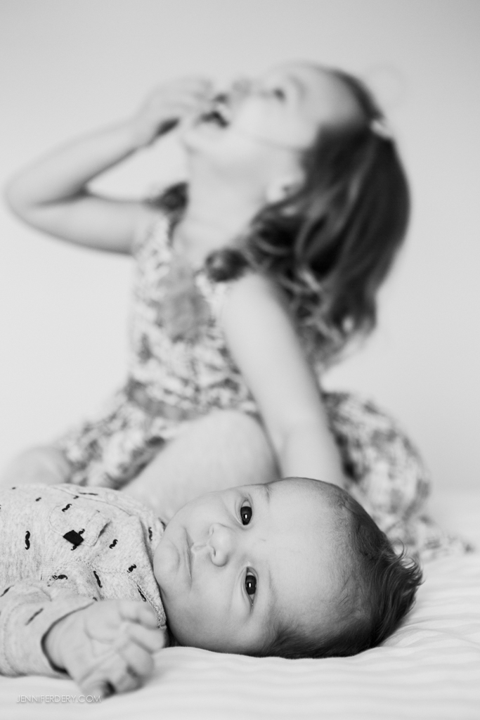 Black and white photo of a baby lying on a bed in the foreground, looking at the camera, with an older child sitting behind, out of focus, laughing and looking upward.