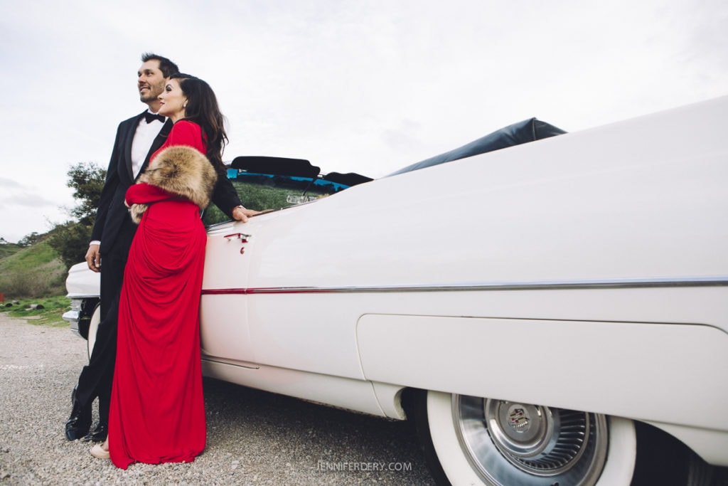 A man in a tuxedo and a woman in a red dress with a fur shawl lean against a classic white convertible. They stand on a gravel path with greenery and trees in the background, under a cloudy sky.