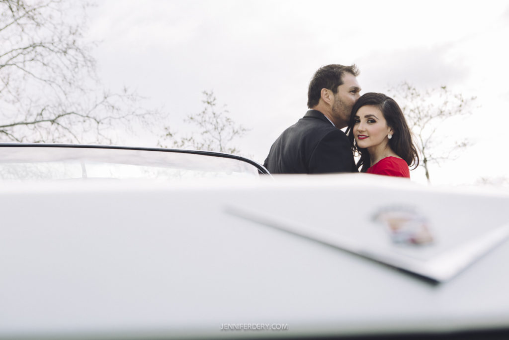 A man in a black suit and a woman in a red dress stand closely together, smiling, with a vintage car in the foreground. Bare trees and a cloudy sky provide the backdrop.
