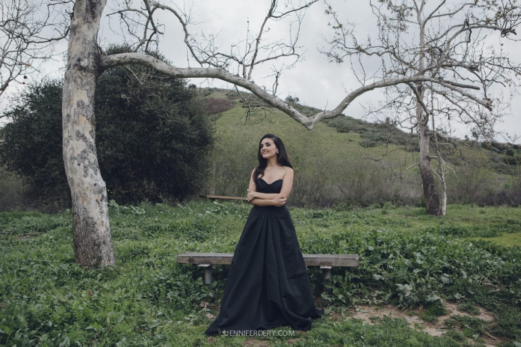 A woman in a long black dress stands in a grassy area with leafless trees and hills in the background. Overcast sky above. She is smiling with arms crossed, leaning against a wooden bench.