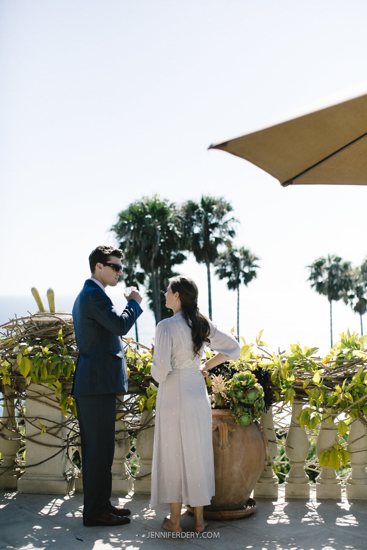 A man in a suit and a woman in a dress stand on a sunlit terrace, engaged in conversation. They overlook an ocean view with palm trees under a clear sky. A large potted plant is nearby, and part of a sunshade is visible above them.