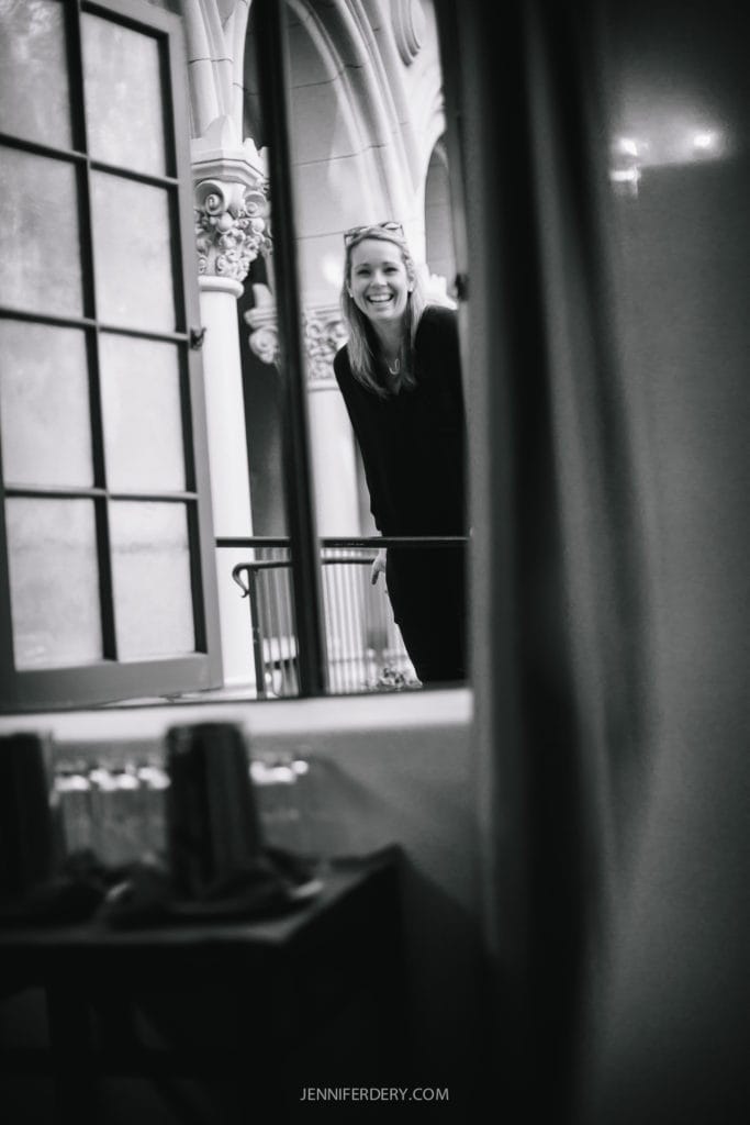A black and white photo captures Ashley of Bliss Events standing and leaning slightly forward. She is visible through a window from inside a room at Balboa Park, with ornate architectural details in the background, perhaps hinting at a charming wedding scene.