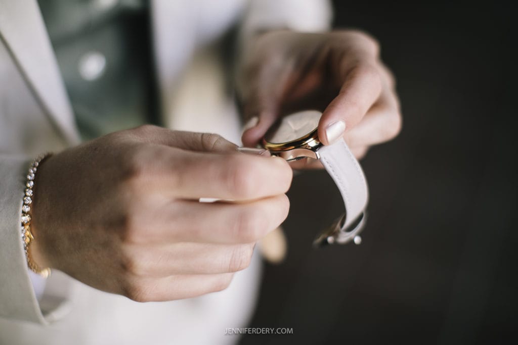A close-up of a person’s hands fastening a white wristwatch with a gold case. The person is wearing a light-colored blazer and a green shirt. A gold bracelet on their wrist adds to the elegance, reminiscent of the sophistication present at a Balboa Park wedding. The image is detailed and focused on the watch.