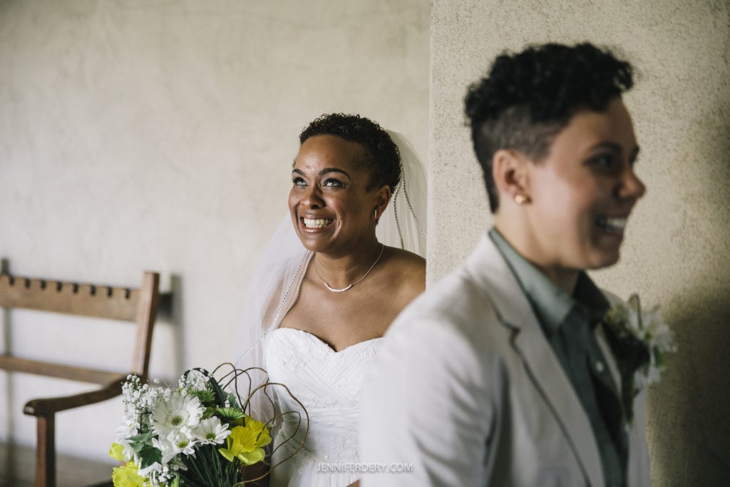 Two people smiling at their Balboa Park wedding. On the left, a person in a white dress holding a bouquet of yellow and white flowers. On the right, another person in a light-colored suit facing away from the camera. Both appear joyful and excited.
