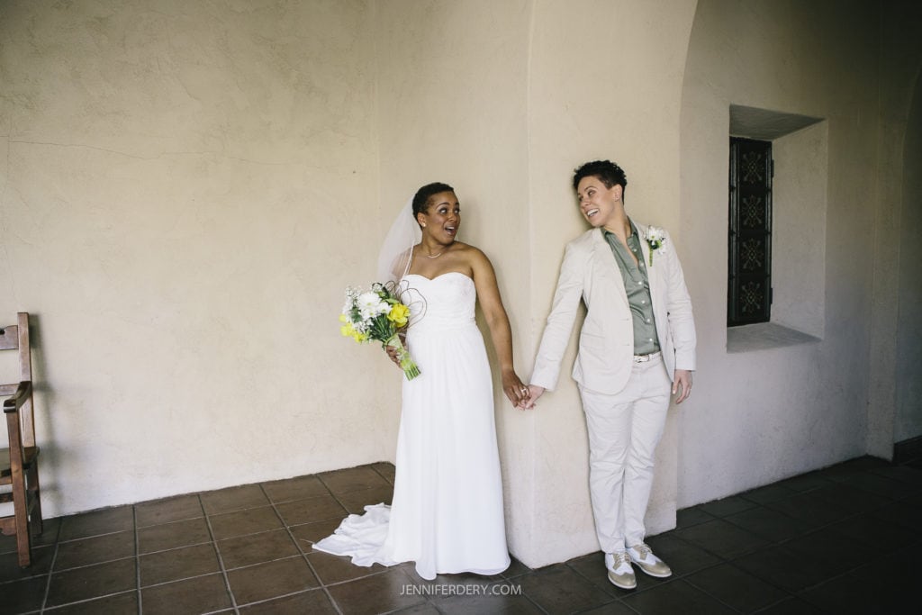 A couple stands hand in hand in an arched hallway at Balboa Park. One person in a strapless white gown, holds a bouquet of flowers and looks at their partner. Her partner, dressed in a beige suit, smiles back at them. A wooden chair is visible in the background.