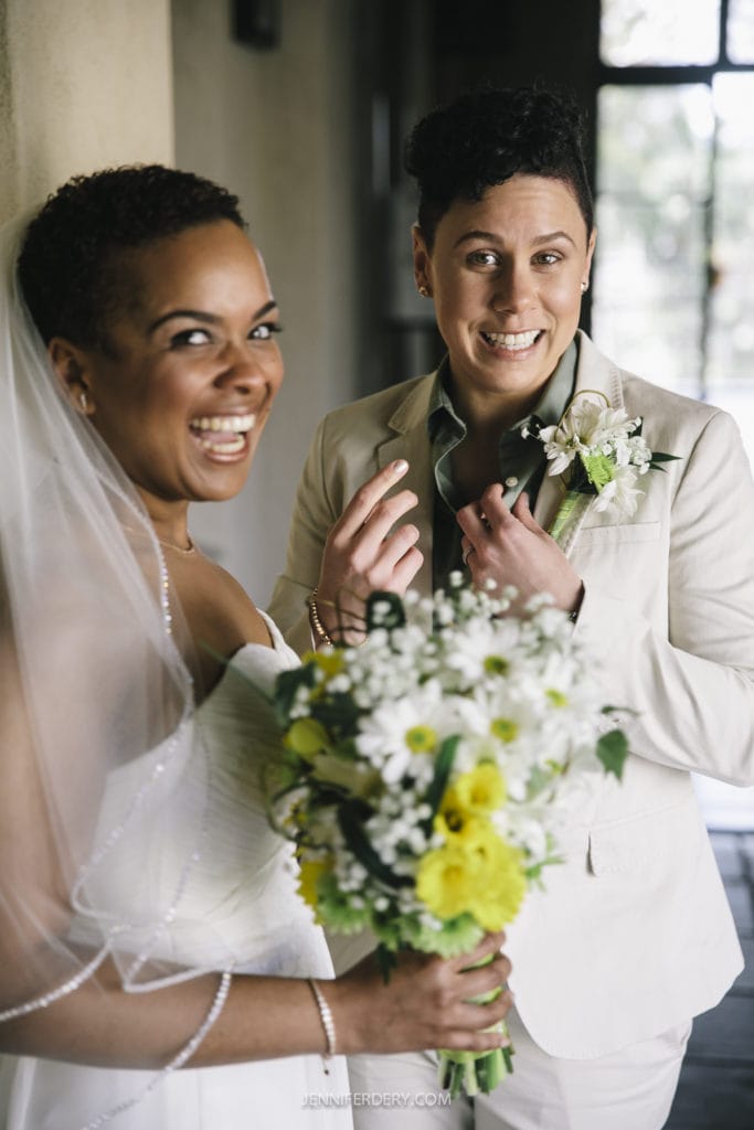 Two people are smiling at the camera on their wedding day in Balboa Park. One is wearing a white wedding dress and holding a bouquet of yellow and white flowers, while the other is in a light-colored suit also holding a small bouquet. Both appear happy and joyful, radiating love in this beautiful setting.