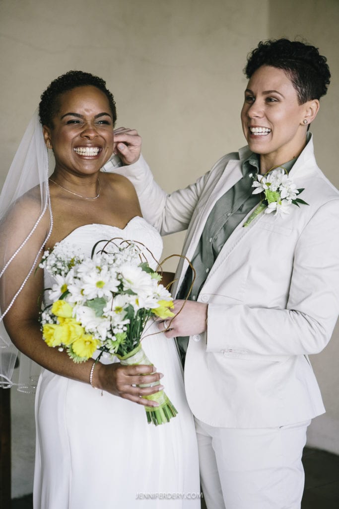 A couple on their wedding day stand together in Balboa Park, both smiling joyfully. One in a white strapless gown holds a bouquet of white and yellow flowers, while the other in a light suit with a boutonniere lightly touches her partner's cheek. Both appear very happy.
