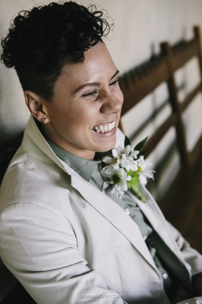 A person with short curly hair and wearing a light beige blazer and a green shirt smiles broadly. A small white boutonnière is pinned to their lapel, suggesting they are at a Balboa Park wedding. They are seated on a wooden bench against a light-colored wall, giving the image a warm and joyful atmosphere.