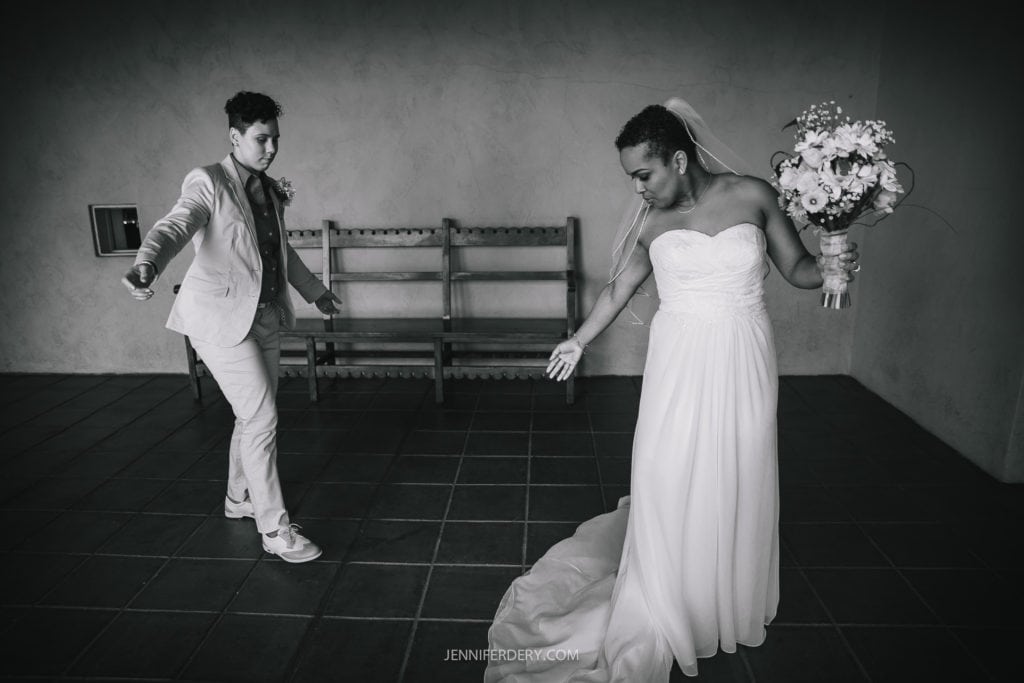 A black and white photo captures a Balboa Park wedding scene. A person in a white bridal gown and veil, holding a bouquet, looks down at their dress. Another person, dressed in a light-colored suit and tie, extends a hand towards the bride. A bench is visible in the background.