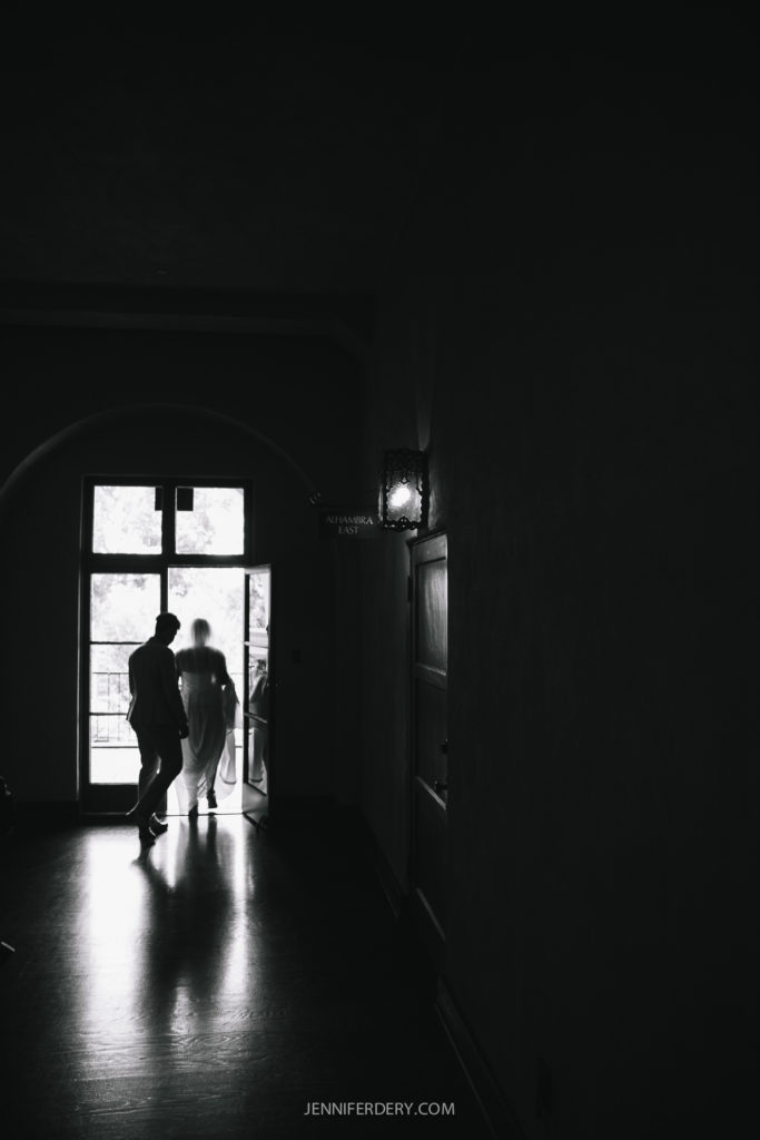 A black and white photo shows a couple standing near an open door, slightly illuminated by outside light. The image captures their silhouettes against the bright outdoor view of Balboa Park, creating a contrasting scene. The floor reflects their figures. The door is arched.