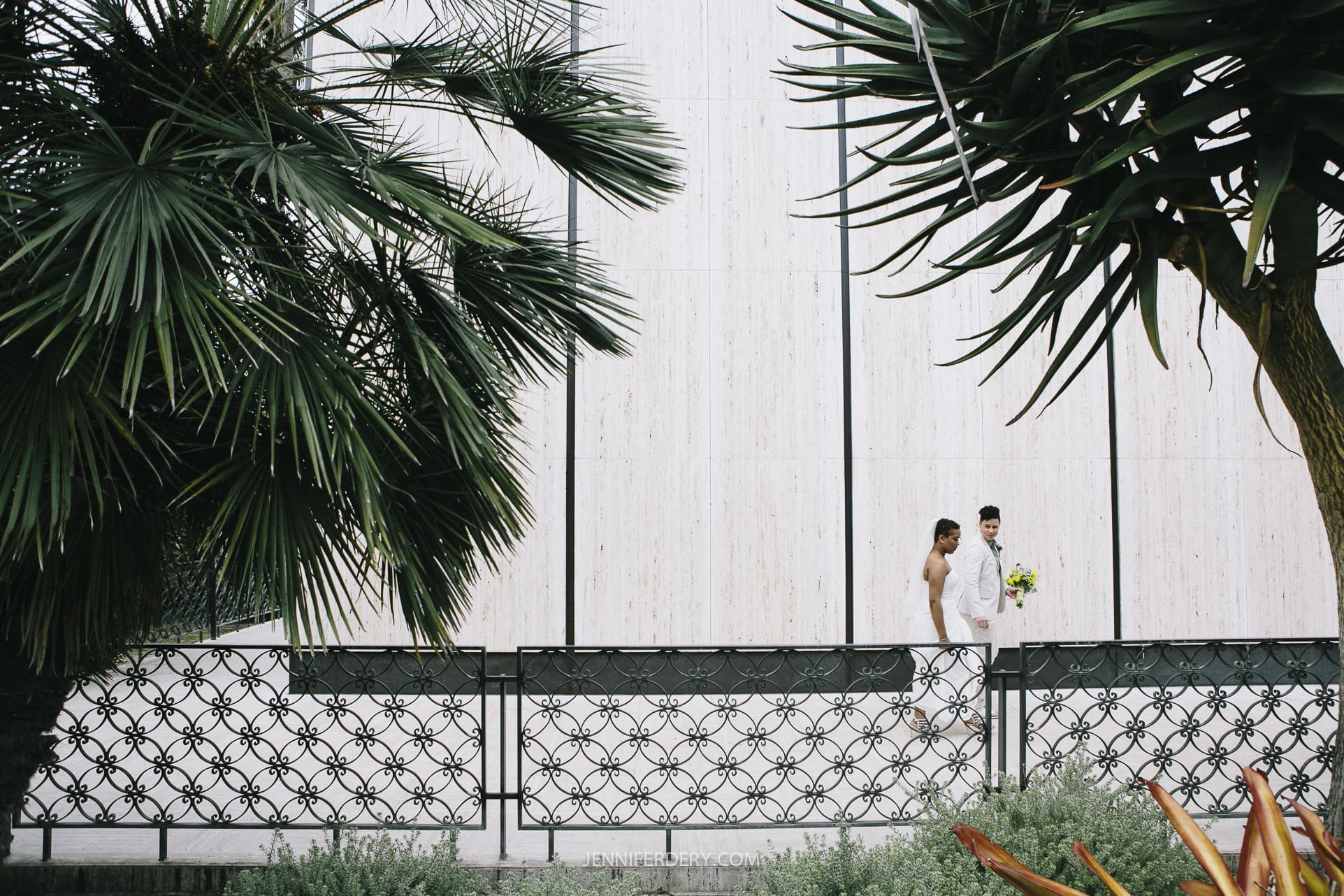 A newlywed couple dressed in white walk alongside a decorative wrought iron fence, flanked by lush green palm trees at Balboa Park. The minimalistic wall background emphasizes the simplicity and elegance of the wedding scene.