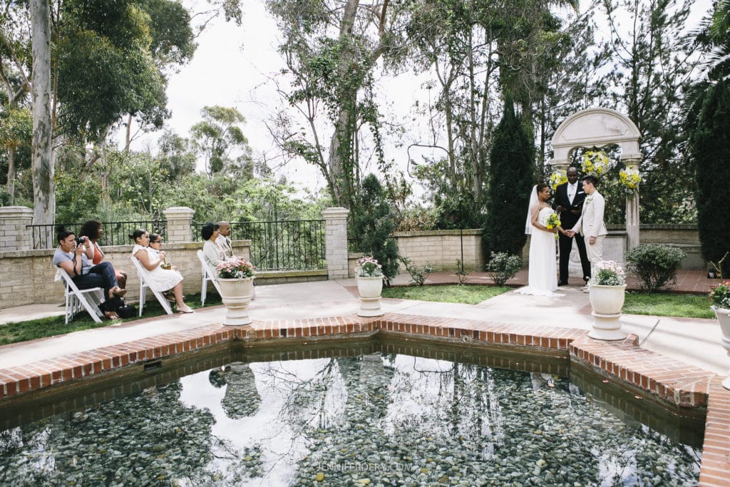 A couple is getting married in an outdoor ceremony by a scenic pond at Balboa Park. They are standing in front of an officiant near a decorated arch. A few guests are seated on white chairs to the left, surrounded by lush greenery and tall trees.