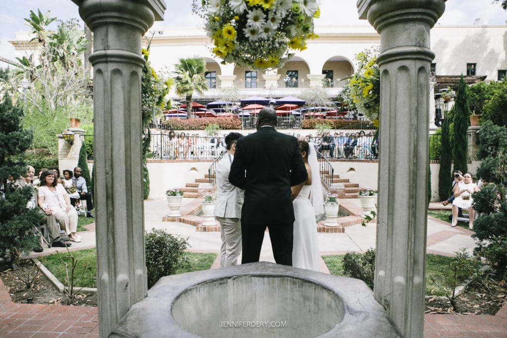 A couple stands before an officiant during an outdoor wedding ceremony at Balboa Park. The officiant holds a microphone, addressing the couple. Guests sit on either side, and a floral arrangement is suspended overhead. The picturesque setting features archways, greenery, and an elevated terrace.