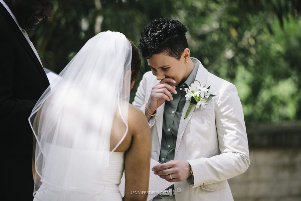 A couple is having an outdoor Balboa Park wedding ceremony. The person on the right, dressed in a light-colored suit and boutonniere, covers their mouth and smiles, appearing emotional. The person on the left, dressed in a white wedding dress and veil, faces away from the camera.