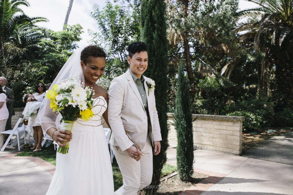 A smiling couple walks hand in hand outdoors during their beautiful Balboa Park wedding. One wears a white wedding dress and holds a bouquet of yellow and white flowers, while the other wears a beige suit. They are surrounded by greenery, and guests can be seen in the background.