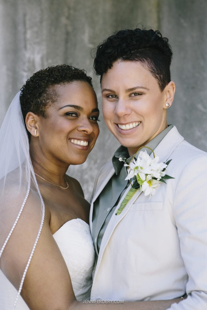 Two smiling women stand closely together at their Balboa Park wedding. One is dressed in a wedding gown with a veil, while the other wears a light-colored suit with a boutonniere. Both are looking at the camera, exuding happiness. The background is a simple, textured surface.