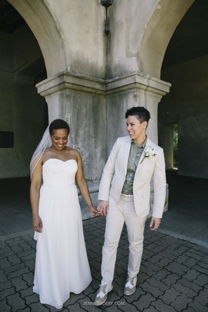A smiling couple, holding hands, stands under an archway in balboa park. One person in a white strapless wedding dress with a veil, the other in a light-colored suit with a white boutonnière. Both are looking at each other lovingly.