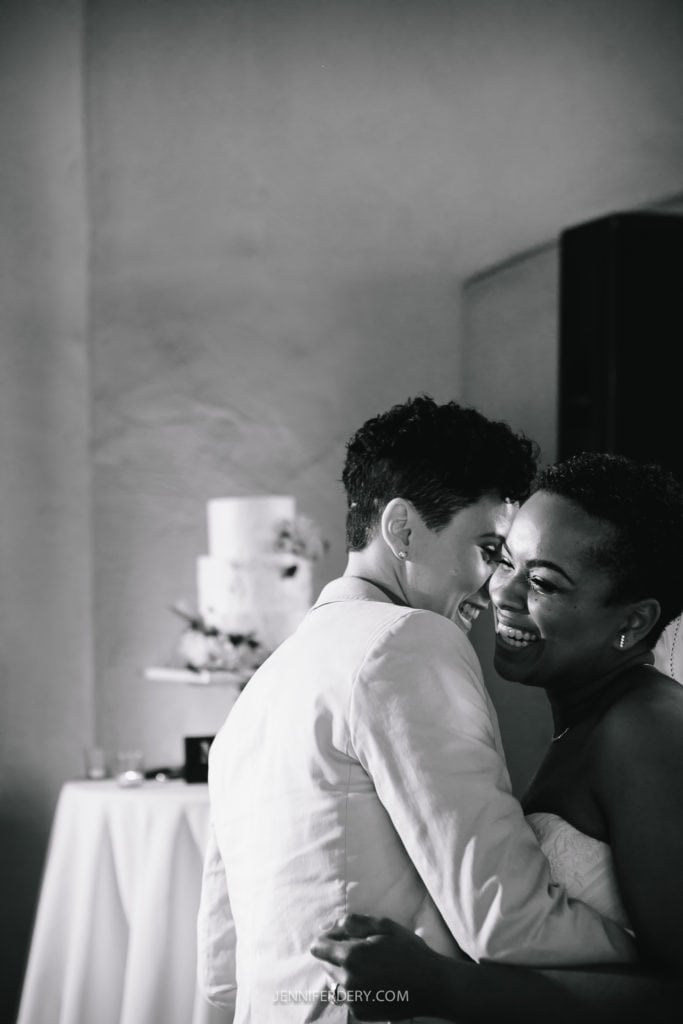 A black and white photo of two people embracing and smiling, at a Balboa Park wedding reception. One is wearing a light-colored suit and the other a strapless dress. They are in front of a table with a tiered wedding cake on it, slightly out of focus in the background.