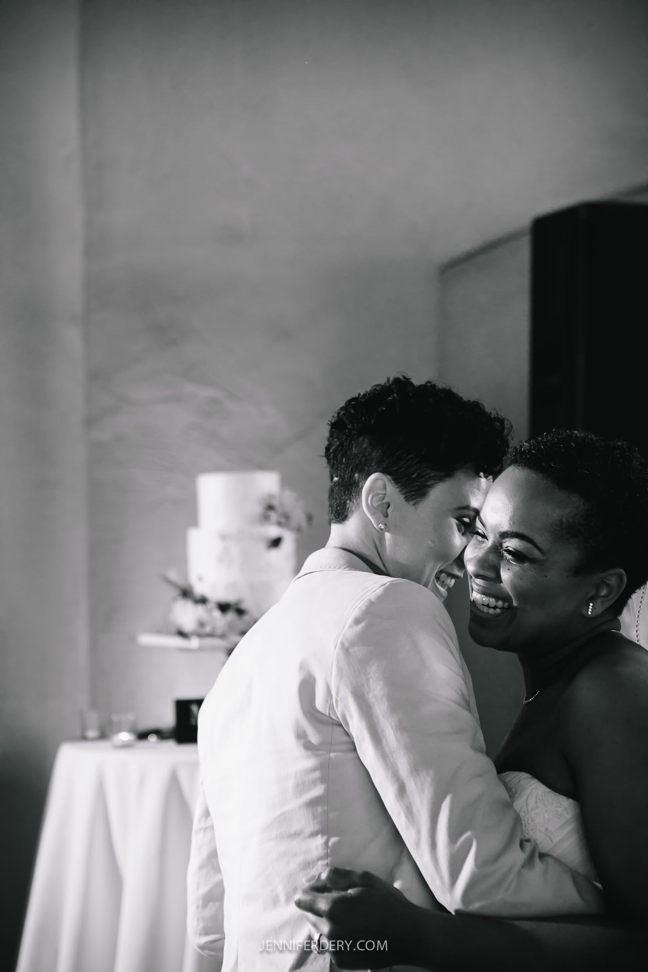 A black and white photo of two people embracing and smiling, at a Balboa Park wedding reception. One is wearing a light-colored suit and the other a strapless dress. They are in front of a table with a tiered wedding cake on it, slightly out of focus in the background. Photographer specializes in Documentary Editorial Wedding Lifestyle 