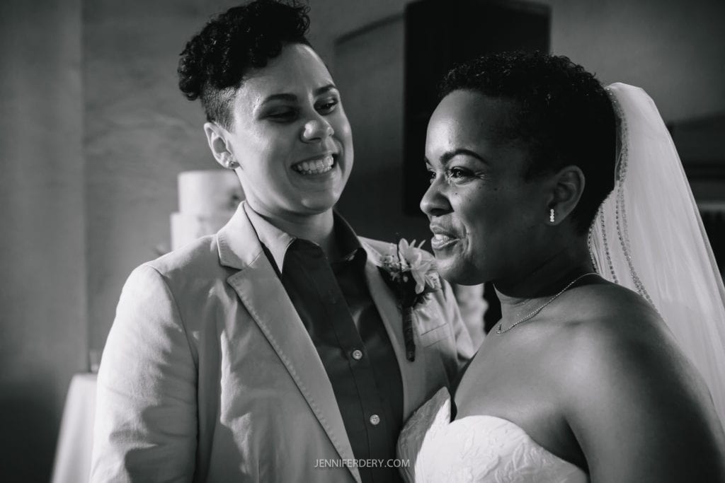 A black and white photo of two newlyweds standing close together. Both are smiling and looking at each other. One person is wearing a light-colored suit with a boutonniere, and the other is wearing a strapless wedding dress with a veil, capturing the elegance of their Balboa Park wedding.