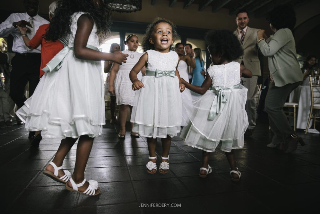 Young children in white dresses with light green sashes are dancing joyfully at a gathering. People in the background, dressed formally, are watching and enjoying the moment. The atmosphere is festive and lively, suggesting a special event at Balboa Park wedding.