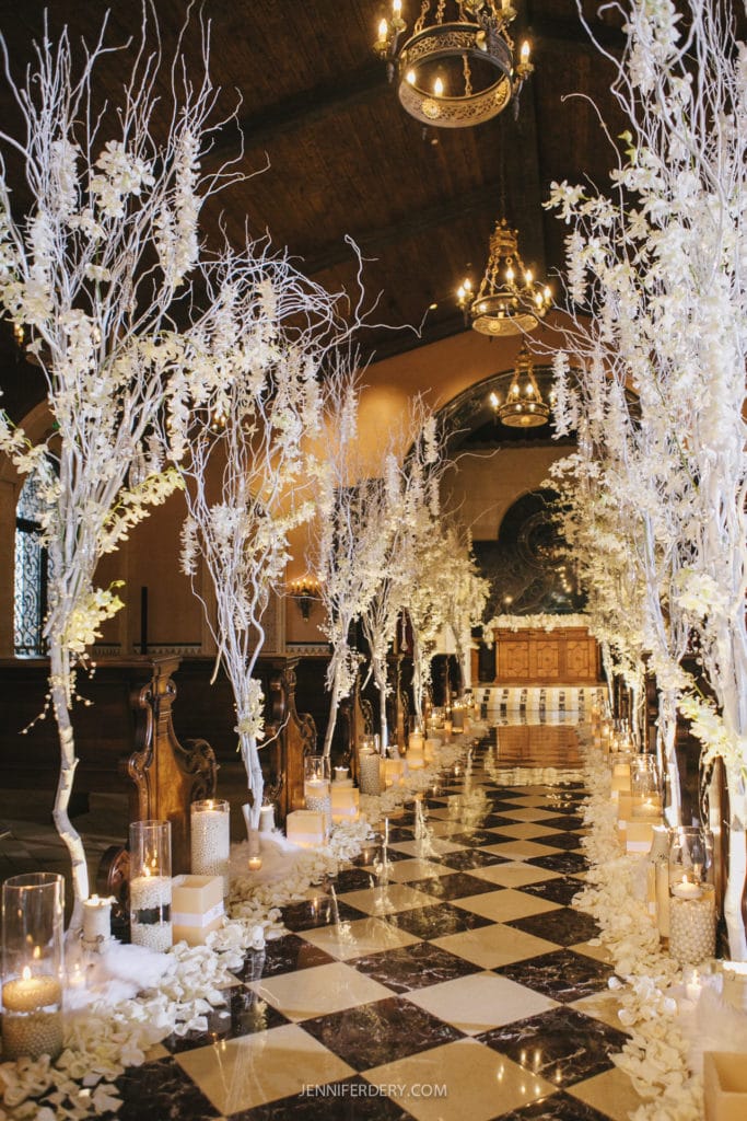 A beautifully decorated wedding aisle with white tree branches and flowers lining the edges. The aisle is illuminated by candles placed in glass holders, creating a warm ambiance. The chapel at the grand del mar features a high arched ceiling with elegant chandeliers.
