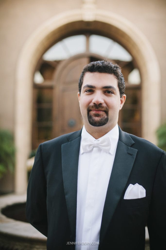 A man with dark hair and a beard is dressed in a black tuxedo with a white dress shirt, whit bow tie, and pocket square. He is standing in front of an ornate wooden door with arched windows. The setting suggests a formal event or wedding.