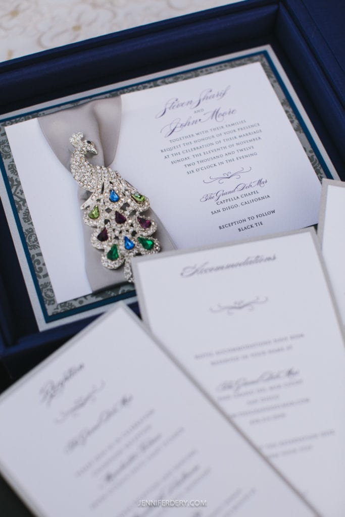 A photo of an elegant wedding invitation set arranged in a navy blue box. The central invitation features a jeweled peacock embellishment and formal text, surrounded by additional cards detailing the event's proceedings. The design has a luxurious and sophisticated feel.