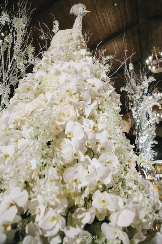A towering floral arrangement adorned with delicate white flowers, including orchids and roses, and topped with a decorative piece resembling a white peacock. The background features elegant, tall floral displays under a high ceiling with wooden beams.