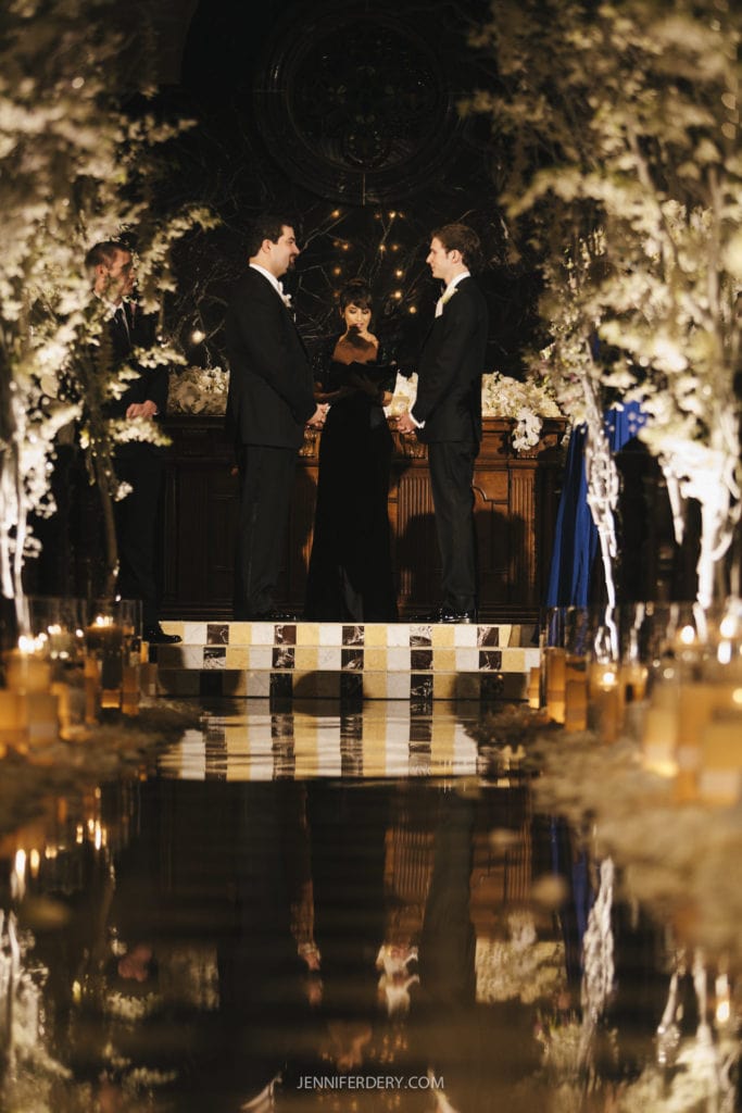 Two grooms, dressed in black suits, stand facing each other on a small stage in the chapel at the grand del mar. A woman in a long black dress stands between them, holding a microphone. The aisle is decorated with white flowers and candles in glass holders. The scene is in a dimly lit, elegant venue.
