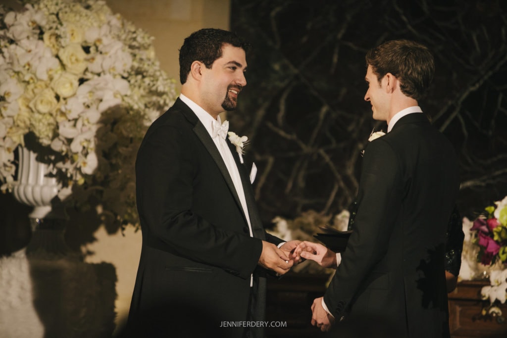 Two men in formal black suits stand facing each other, smiling and holding hands, during a wedding ceremony. One man is holding a piece of paper, possibly vows. A large floral arrangement is visible in the background along with a dark marble wall.